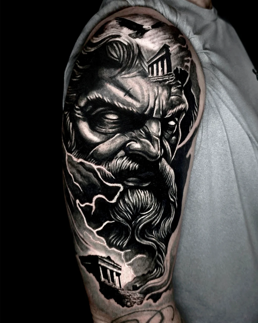 A detailed black and gray tattoo on an upper arm depicts a stern, bearded mans face surrounded by storm clouds and ancient Greek temple ruins, creating a dramatic and mythological scene.