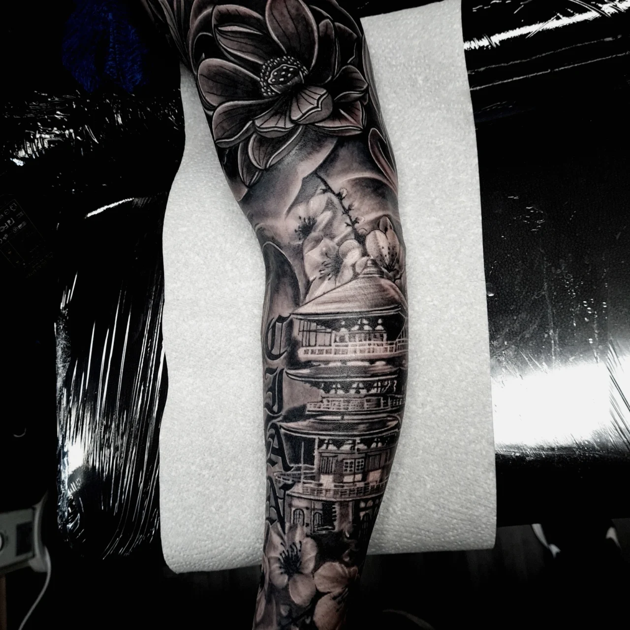 A detailed black and grey tattoo sleeve featuring a large lotus flower, pagodas, and mountains covers a persons forearm, resting on a padded white surface.