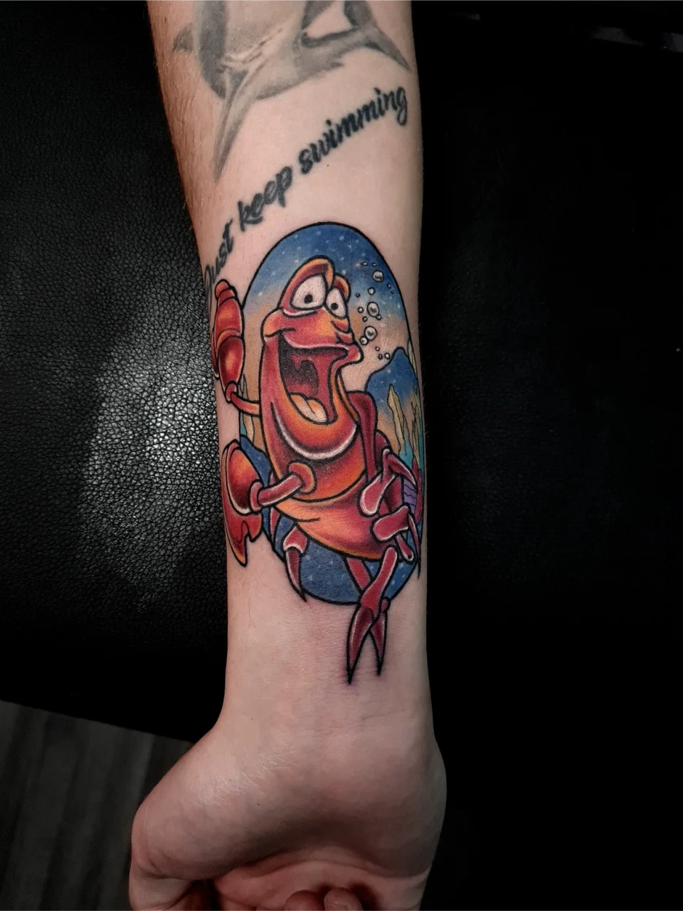 A colorful tattoo of a smiling cartoon crab inside an oval, with bubbles and an underwater background, on a person’s forearm. Part of a “Just keep swimming” tattoo is visible above.
