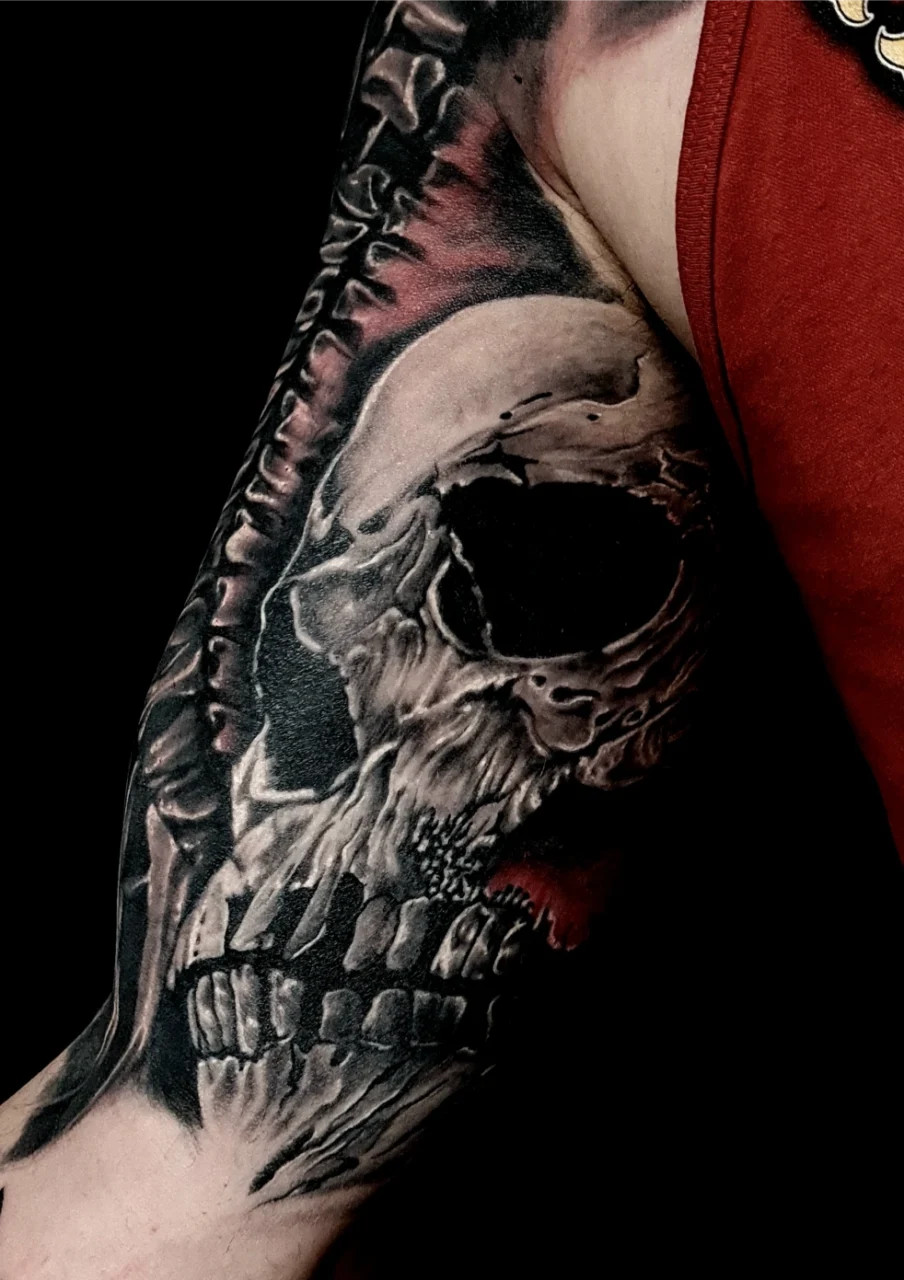 A detailed black and gray skull tattoo is inked on a persons upper arm, blending realistic shading and highlights. The person is wearing a sleeveless red top, exposing the tattooed arm against a black background.