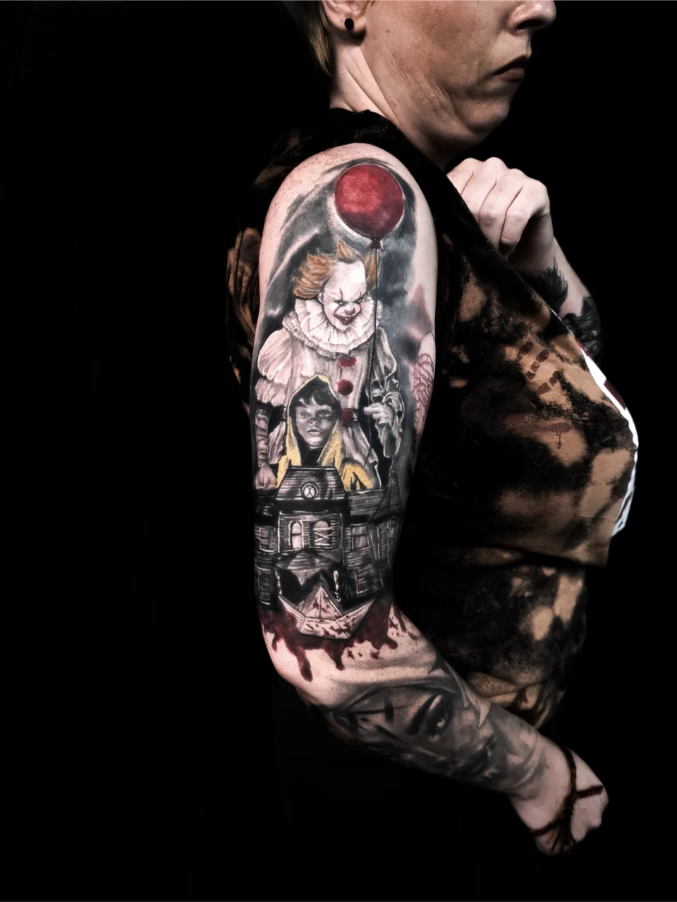 A person’s upper arm displays a detailed tattoo featuring characters and elements from the horror film IT, including Pennywise the clown holding a red balloon and a child in a yellow raincoat, against a dark background.