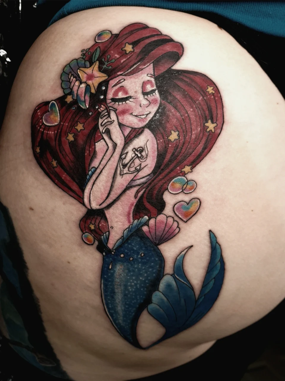 Colorful tattoo of a smiling mermaid with long red hair, blue tail, and star accents. She is surrounded by seashells, bubbles, and hearts, with an anchor tattoo on her arm.