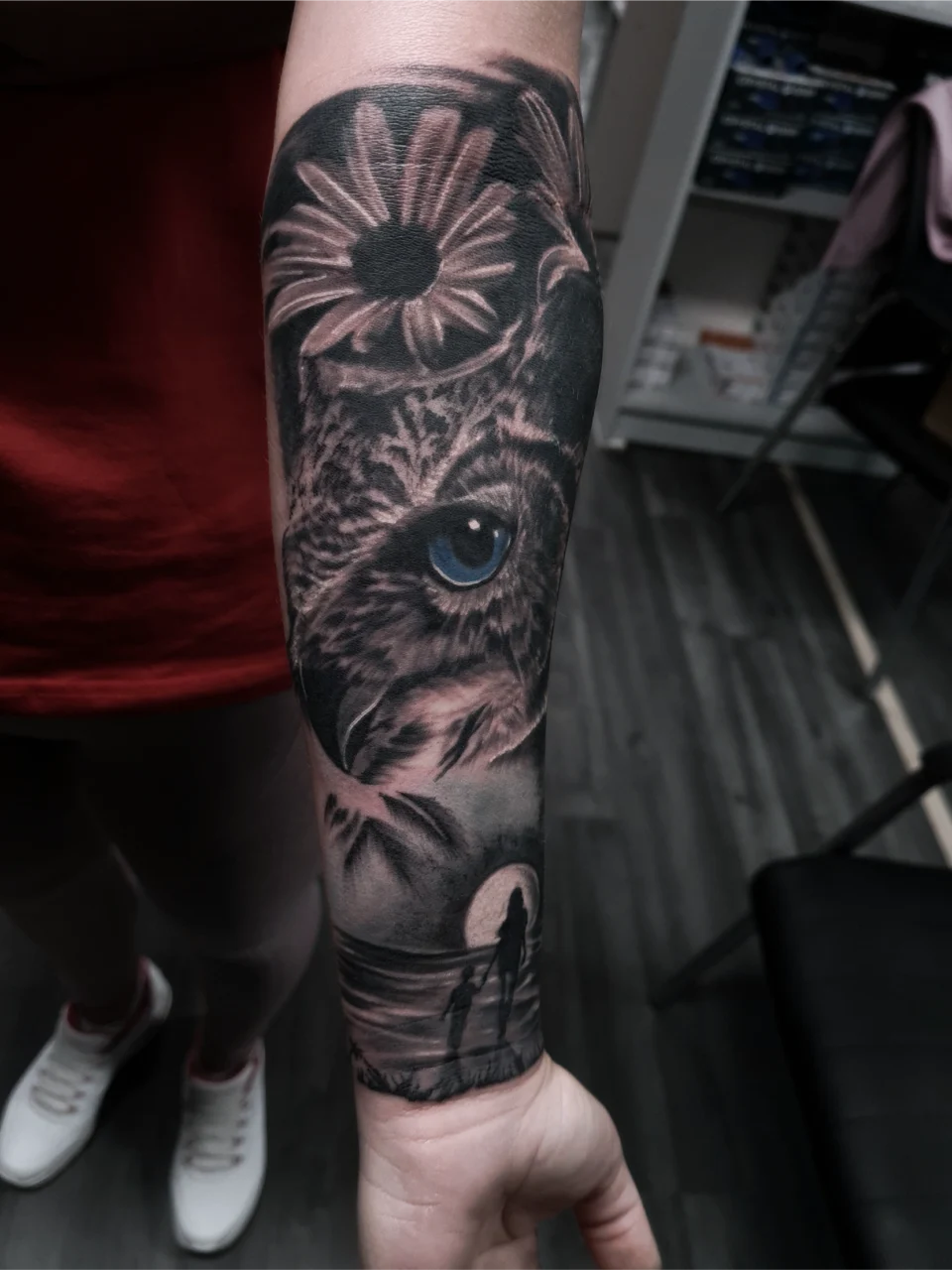 A realistic black and gray forearm tattoo featuring an owl with a blue eye, a daisy above its head, and a silhouette of two people walking on a beach at sunset.