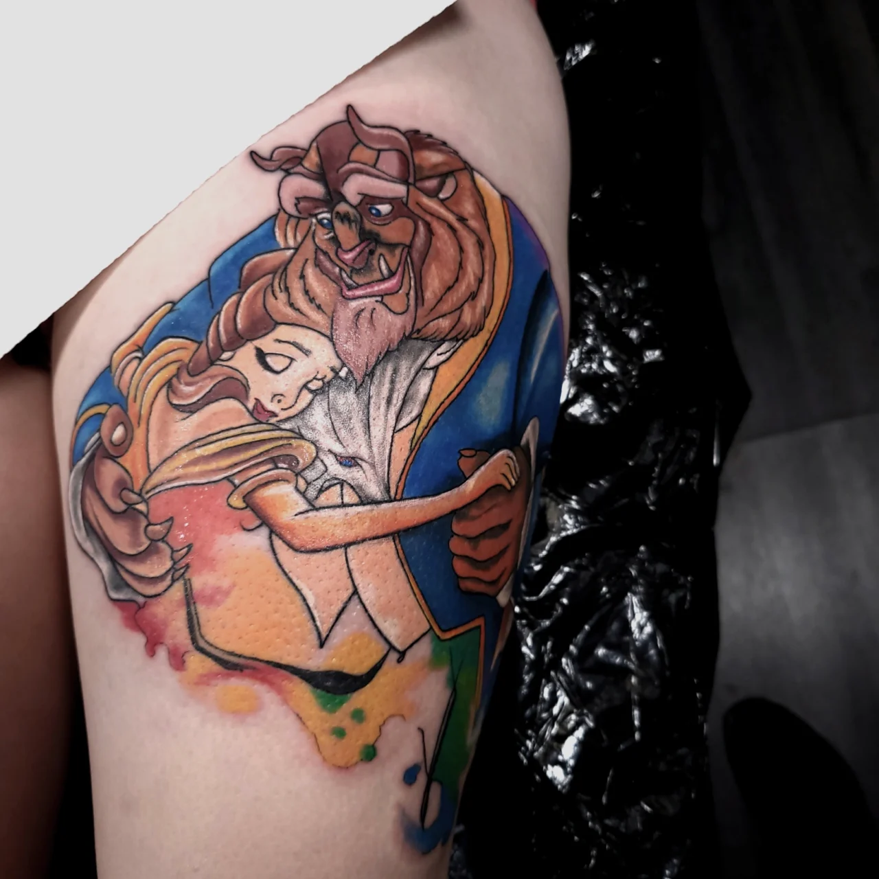 A colorful tattoo on a persons thigh depicts Belle and the Beast from Beauty and the Beast dancing together, with vibrant watercolor-style accents surrounding the characters.
