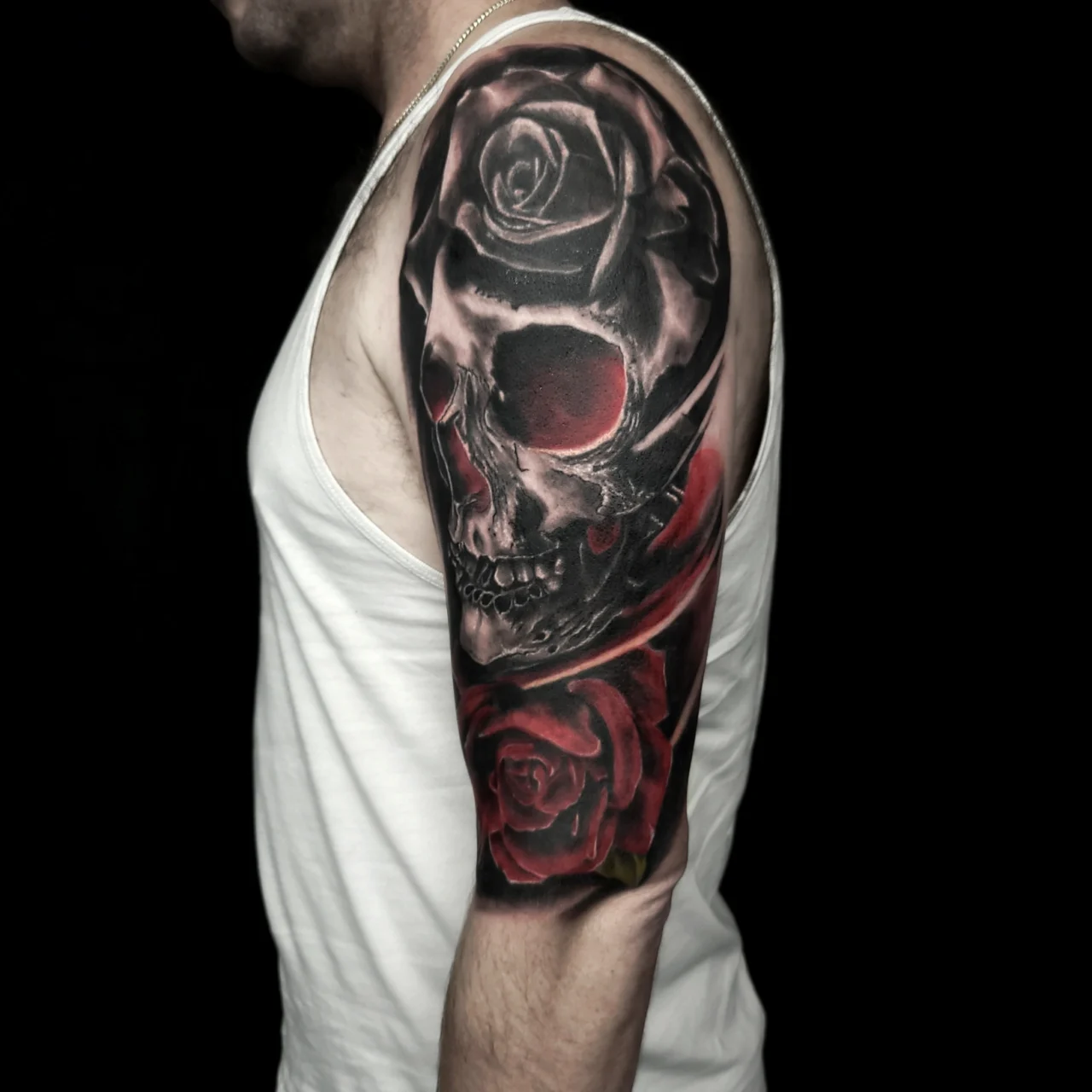 A man in a white tank top displays a detailed upper arm tattoo featuring a realistic black and gray skull surrounded by dark and red roses on a black background.