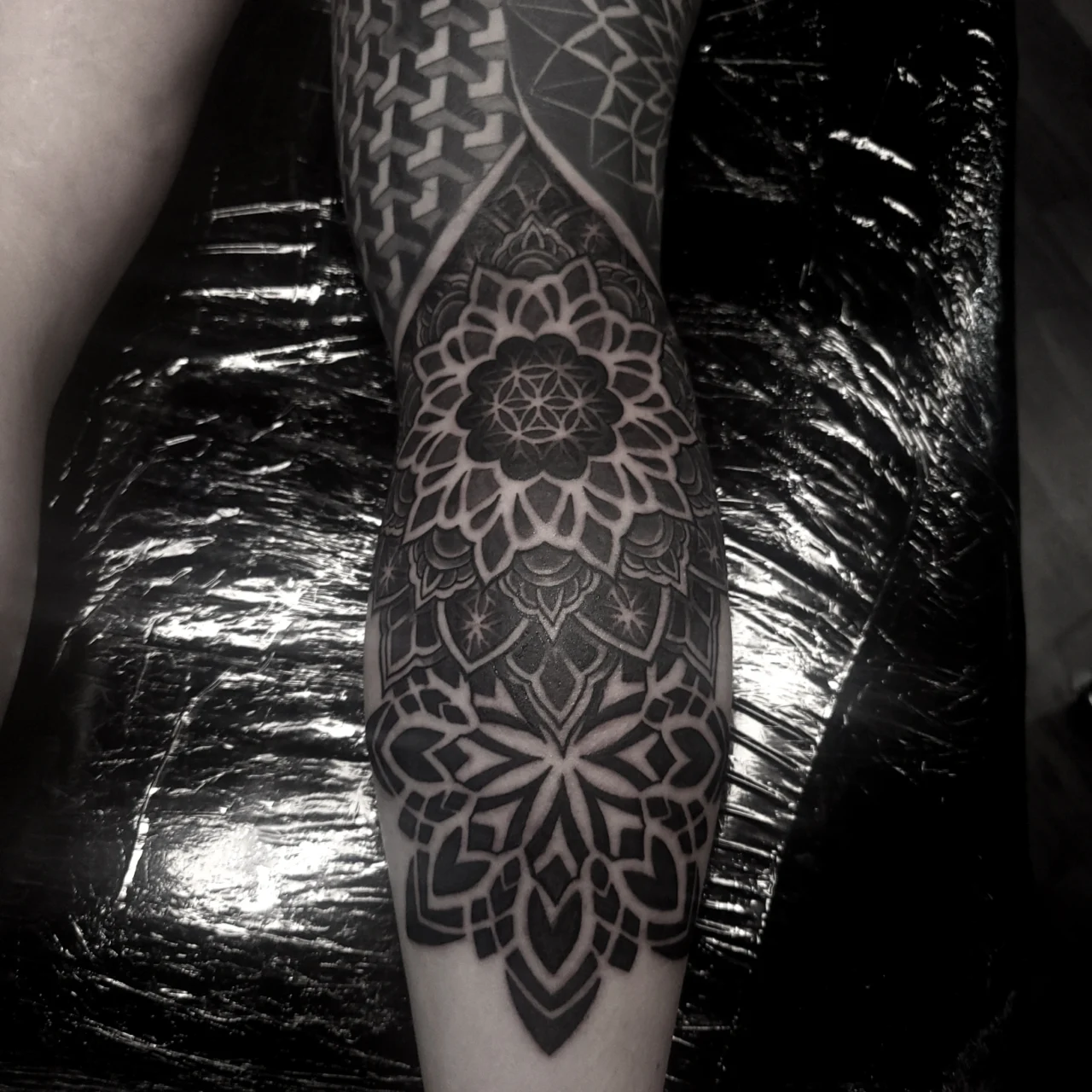 A black and grey geometric mandala tattoo covers a persons knee and thigh, featuring intricate symmetrical patterns and bold linework. The tattoo design blends floral and abstract elements.