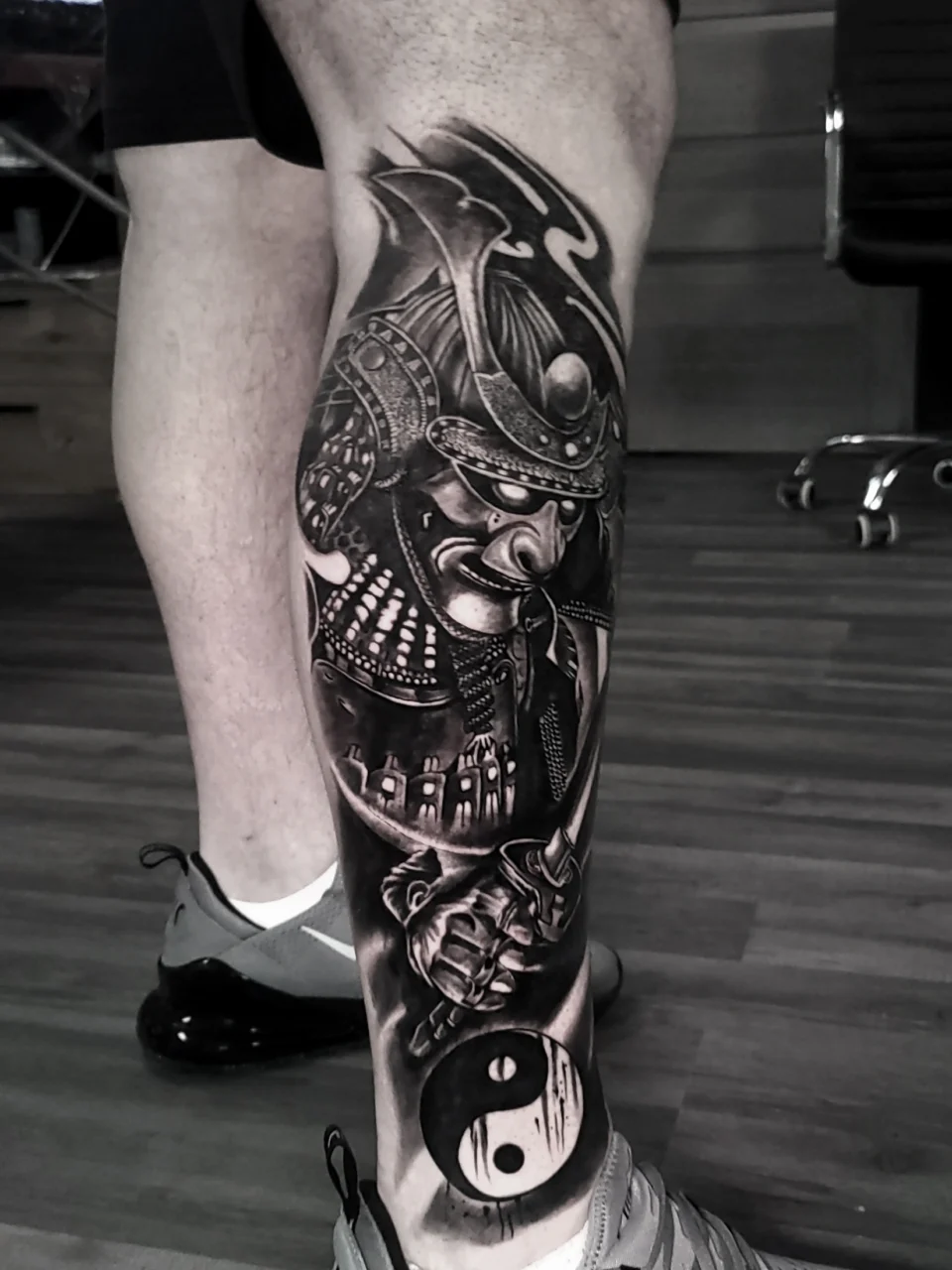 A detailed black and grey tattoo of a samurai warrior holding a sword, covering the entire calf. At the bottom of the tattoo is a yin-yang symbol. The person is standing indoors, wearing gray shoes.