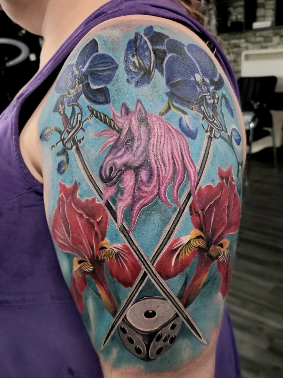 A colorful tattoo on an upper arm features a pink unicorn head, two crossed swords, two purple flowers, two red flowers, and a six-sided die, all set against a blue background.