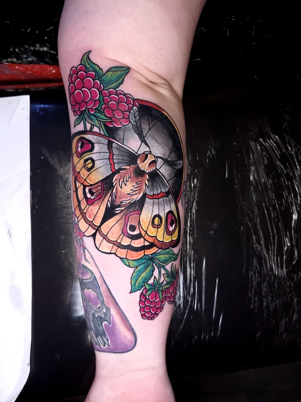 A colorful tattoo on a persons forearm features a detailed butterfly with orange and yellow wings, surrounded by pink berries and green leaves.