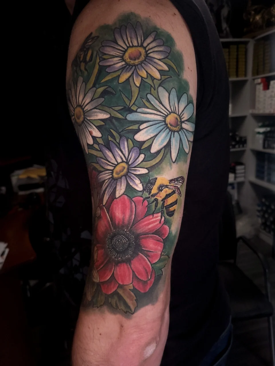 A colorful tattoo on an upper arm features white and purple daisies, a large red flower, green leaves, and a yellow and black bee, all set against a green background.