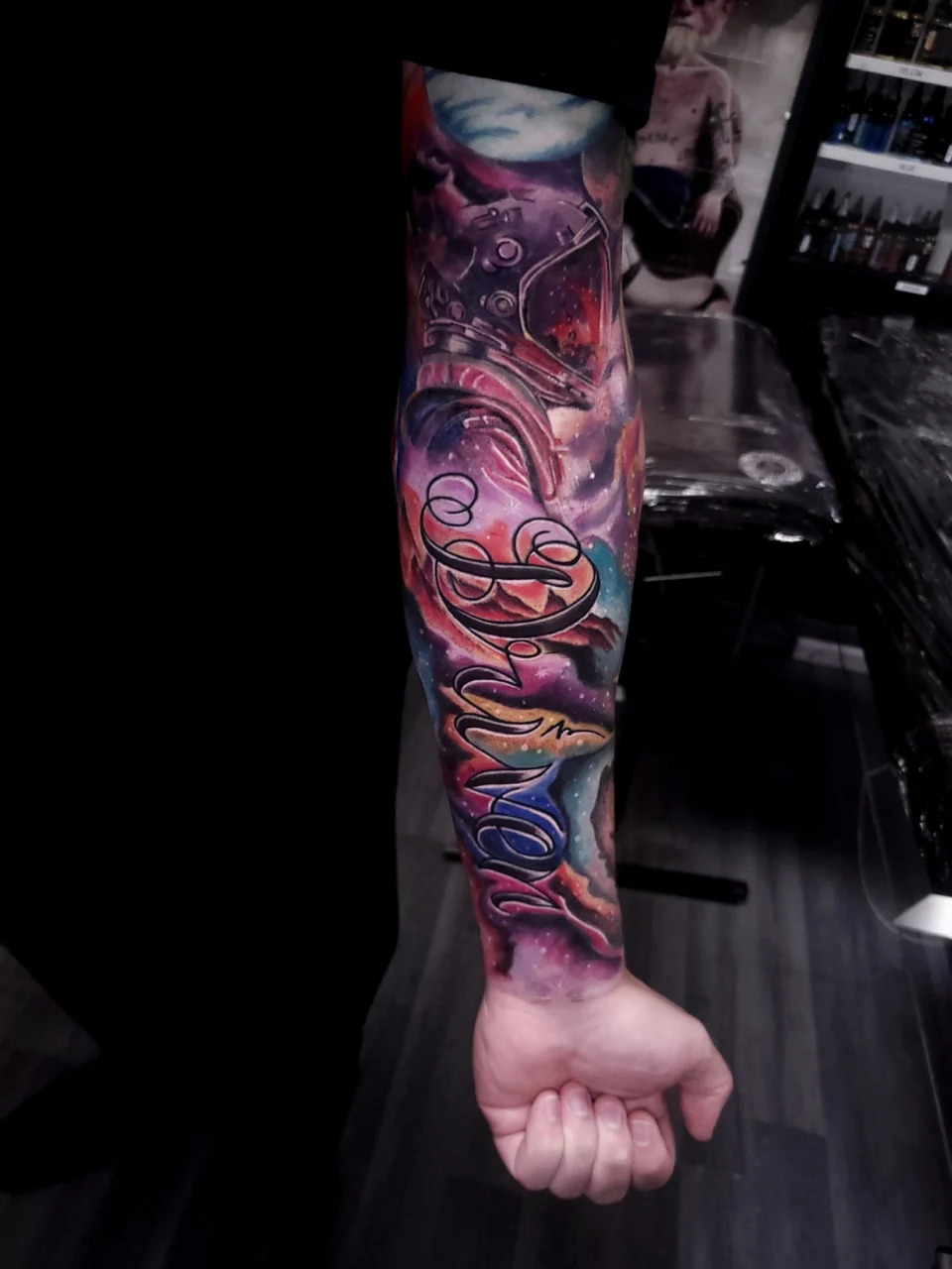 A forearm tattoo featuring vibrant, colorful cosmic imagery with planets and stars, and the word Shine in elegant script across the center. The person’s arm is extended, showing the tattoo’s detail.