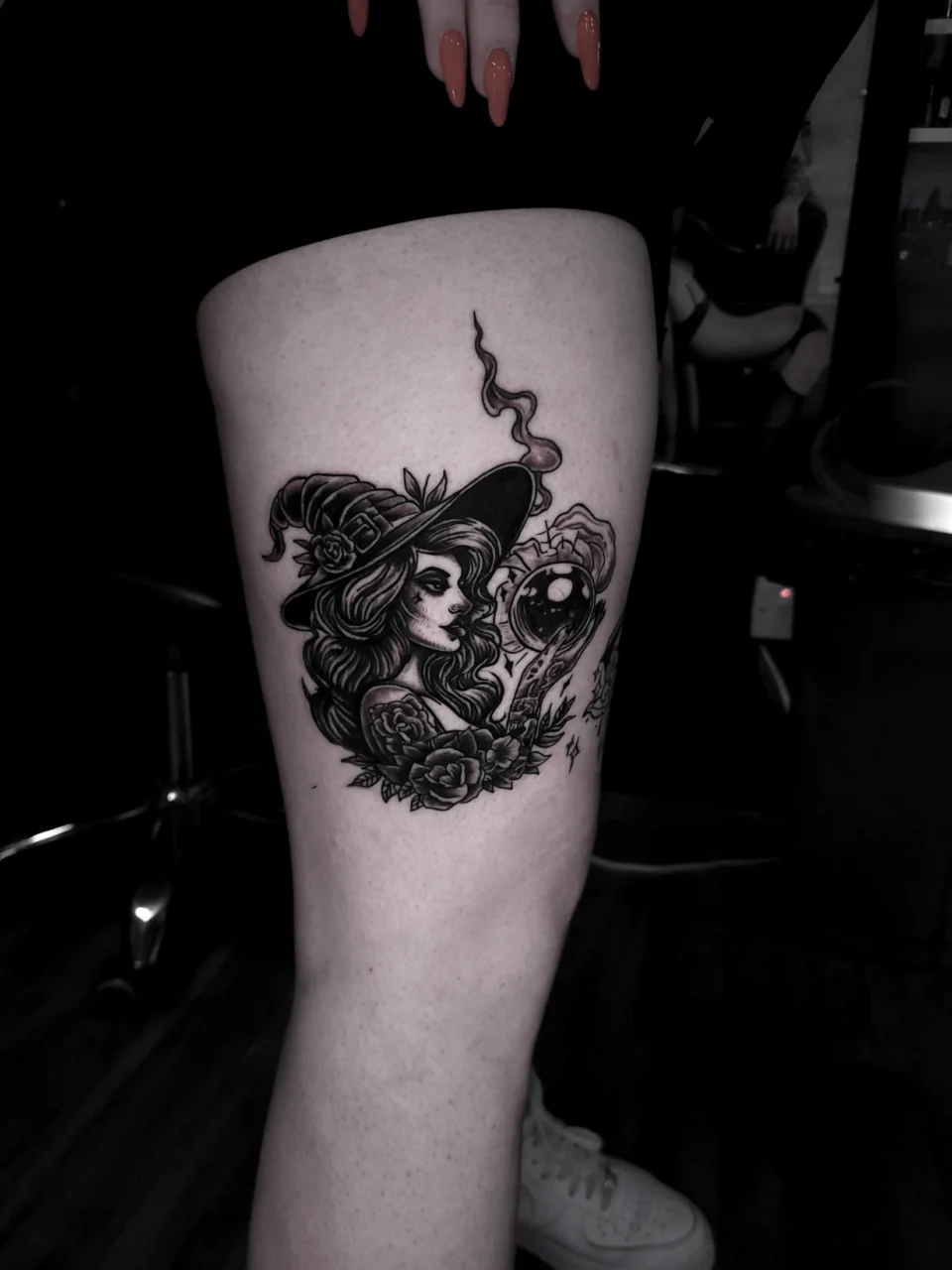 Black and grey tattoo on a persons thigh depicting a witch in a hat surrounded by roses, with a smoking crystal ball and mystical accents. The person has long nails and is standing indoors.
