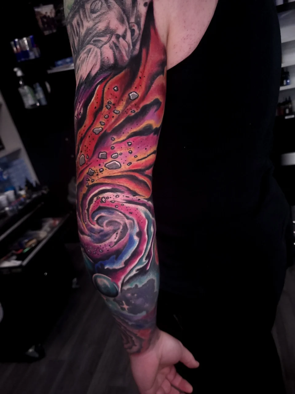 A colorful full-sleeve tattoo featuring a cosmic scene with swirling galaxies, vibrant red, orange, and purple hues, floating rocks, planets, and the face of a large cat at the top, on a persons arm.