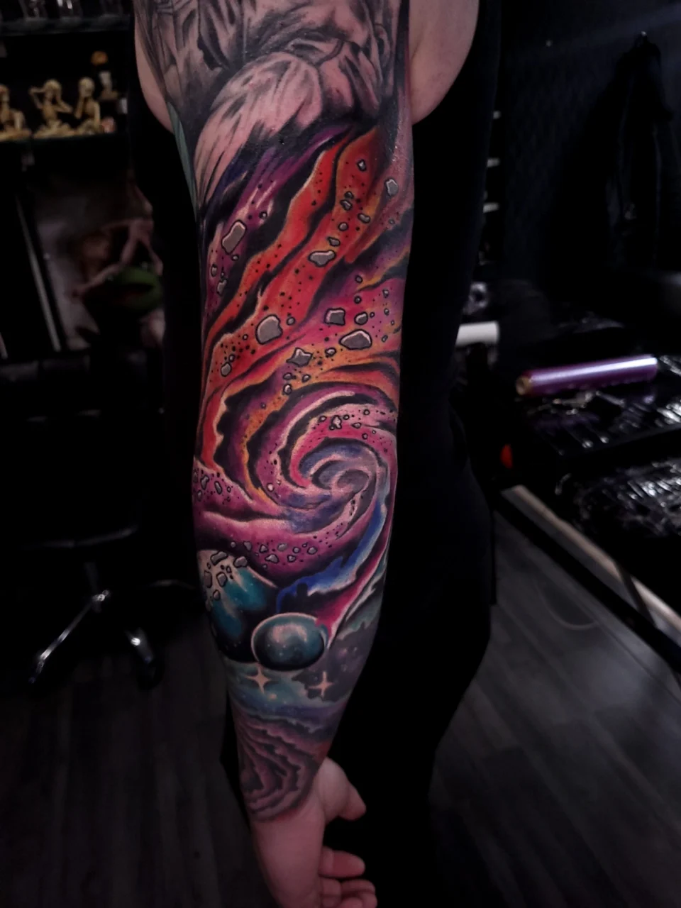 A colorful space-themed tattoo featuring a swirling galaxy, stars, and planets covers a persons forearm, shown in a dimly lit tattoo studio.