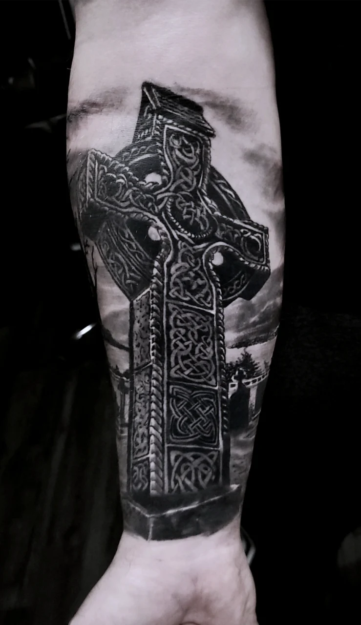 A realistic black and grey tattoo of a Celtic cross with intricate knotwork designs, placed on a persons forearm. The background features cloudy skies and faint silhouettes of stone structures.