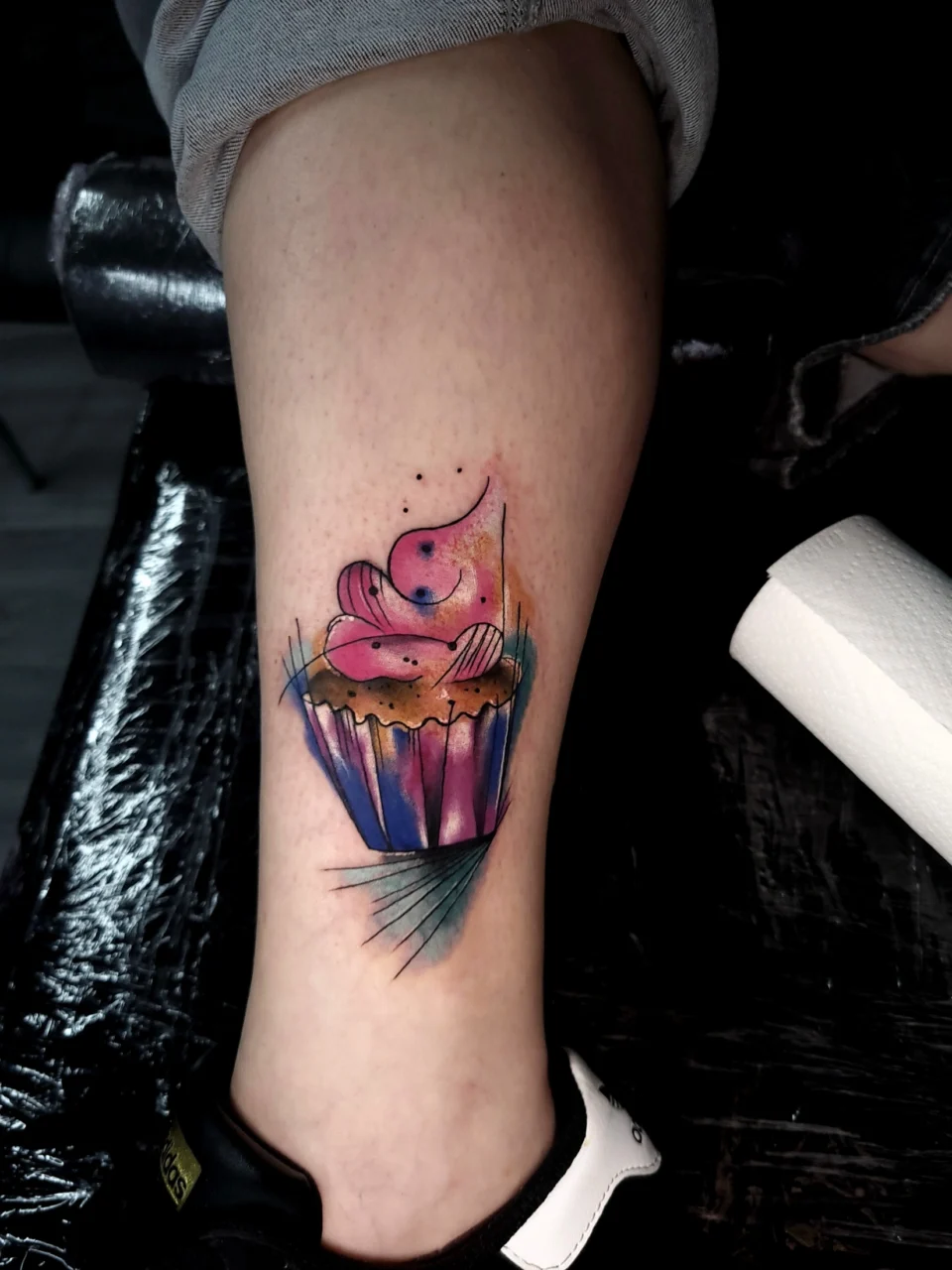 A colorful watercolor-style cupcake tattoo with purple, blue, pink, and yellow hues is inked on a persons calf. The tattoo features artistic splashes and abstract lines.