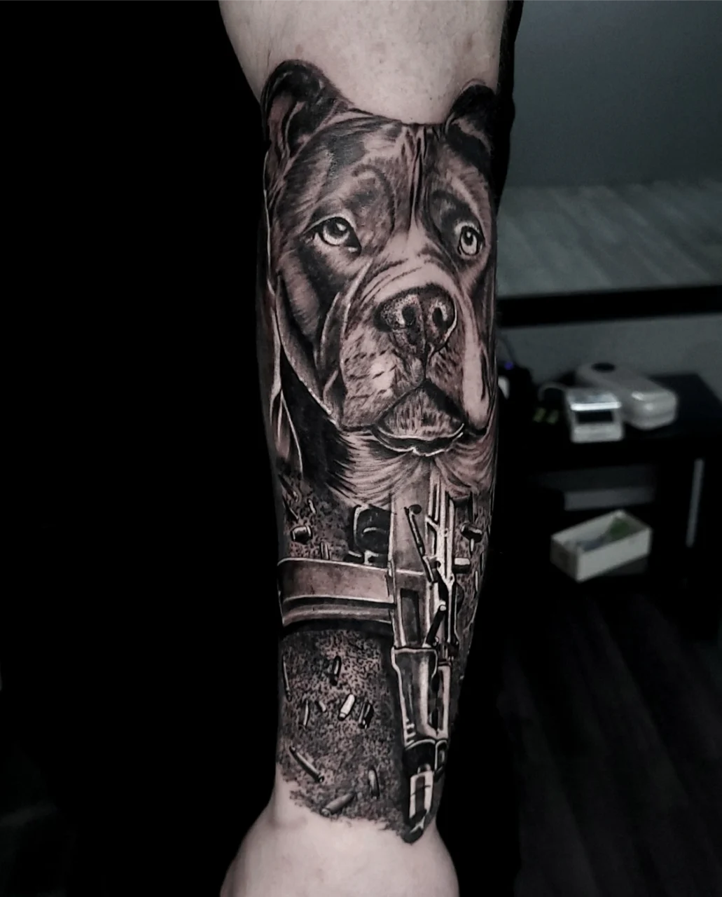 A realistic black and grey tattoo on a forearm features a detailed dog’s face above a scene with construction tools and equipment. The background is indoors, showing part of a table and some items.