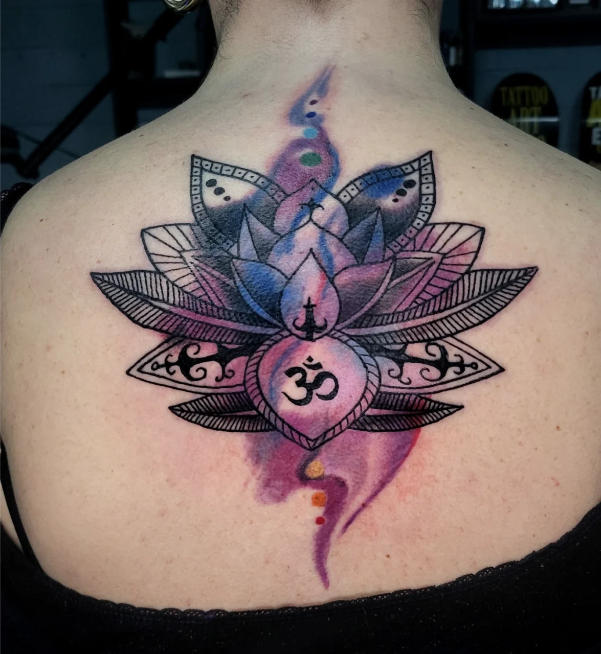 A colorful tattoo of a lotus flower with intricate black outlines and an Om symbol in the center, placed on the upper back. There are small colored chakra dots in a vertical line above and below the lotus.