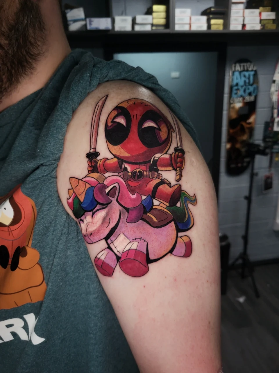 A colorful tattoo on a persons upper arm depicts a chibi-style Deadpool holding two swords while riding a rainbow-maned unicorn. The surrounding setting appears to be a tattoo studio.
