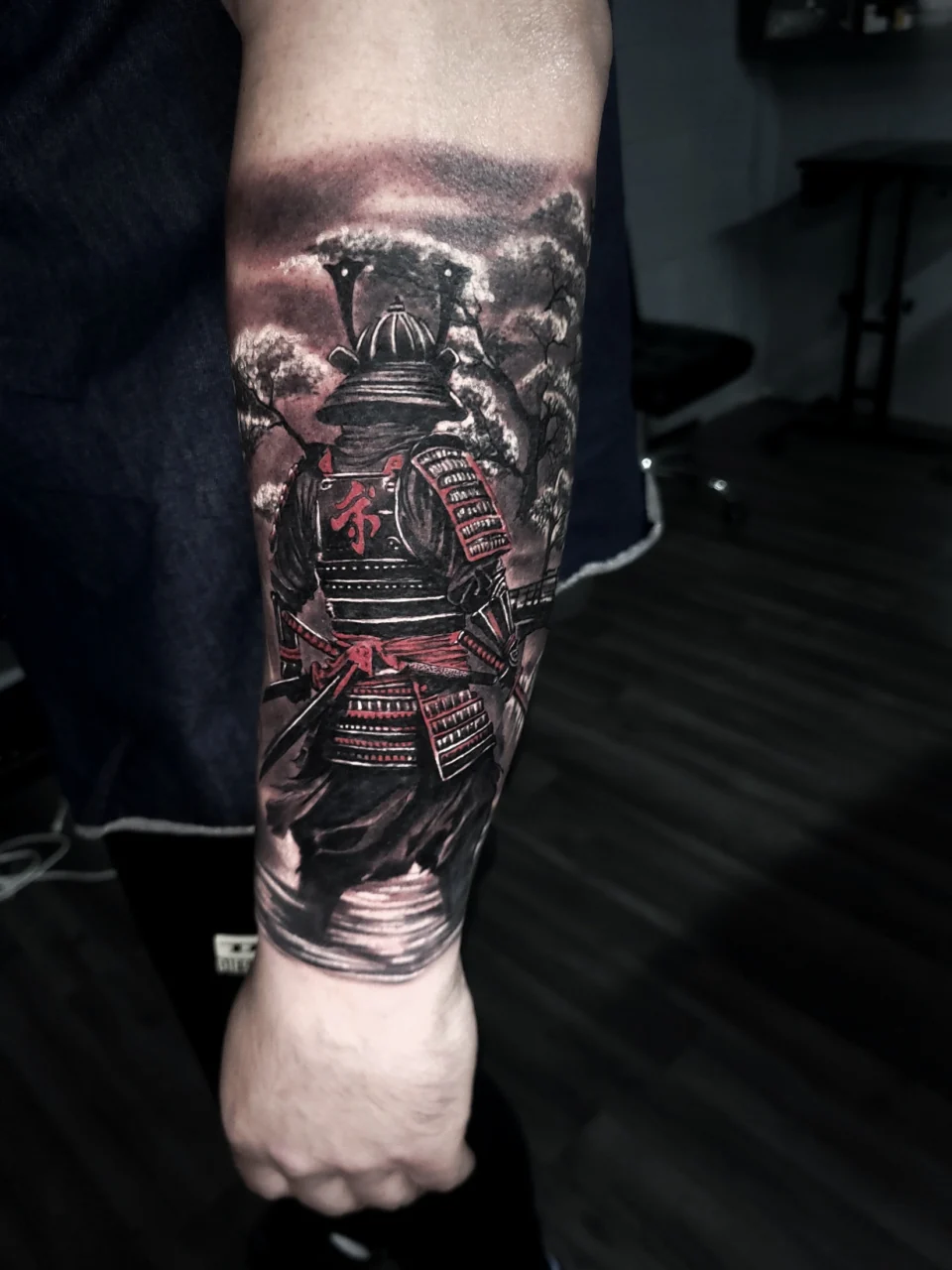 A detailed forearm tattoo of a samurai in armor, facing away, with red accents on the armor, set against a cloudy background. The tattoo is on a persons inner forearm, and the photo has a dark, moody atmosphere.