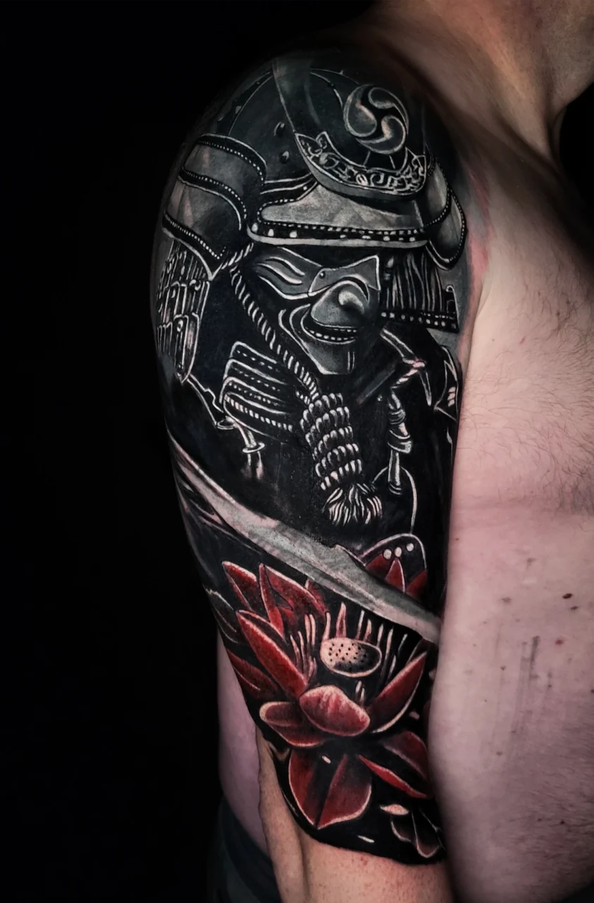 A detailed black and gray samurai tattoo covers an upper arm and shoulder, featuring armor, a helmet, and a red lotus flower with shaded petals near the bicep. The background is dark, highlighting the intricate design.