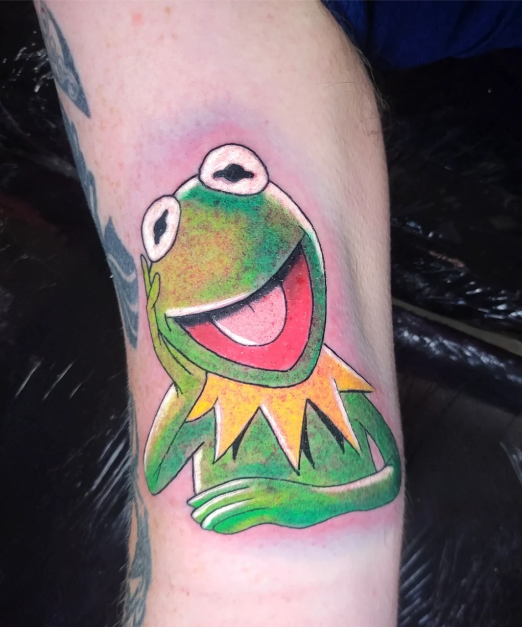 A colorful tattoo of Kermit the Frog, smiling with his head resting on one hand, is inked on a persons inner arm. The tattoo features bright green, yellow, and pink hues.