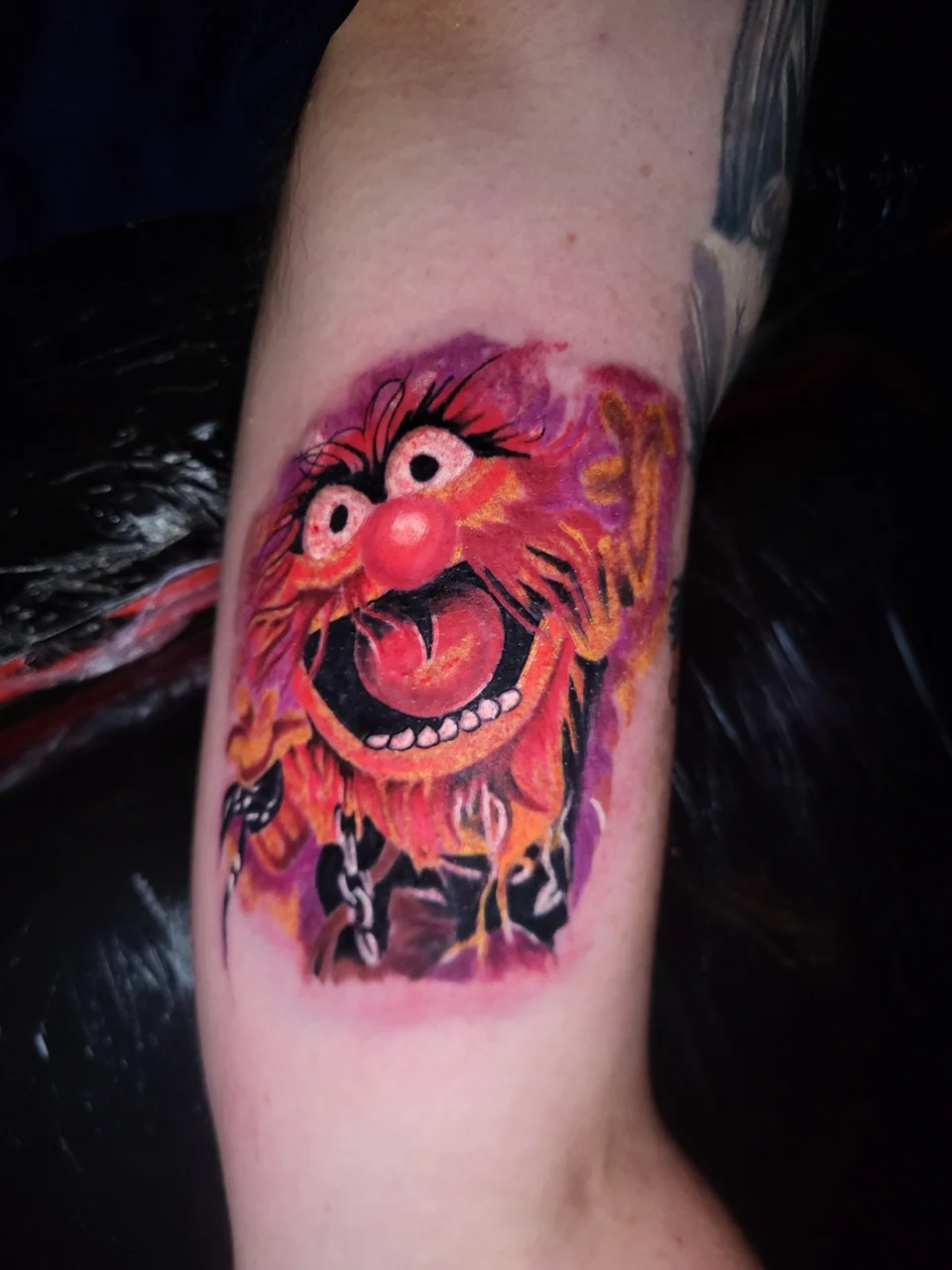 A colorful tattoo of Animal, the wild drummer from The Muppets, featuring his shaggy orange hair, wide eyes, open mouth, and vibrant red and pink background, displayed on a persons arm.