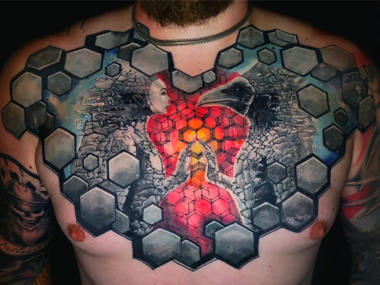 A colorful chest tattoo features a red heart at the center, framed by a woman and a raven facing each other, surrounded by gray hexagonal patterns across the chest and shoulders.