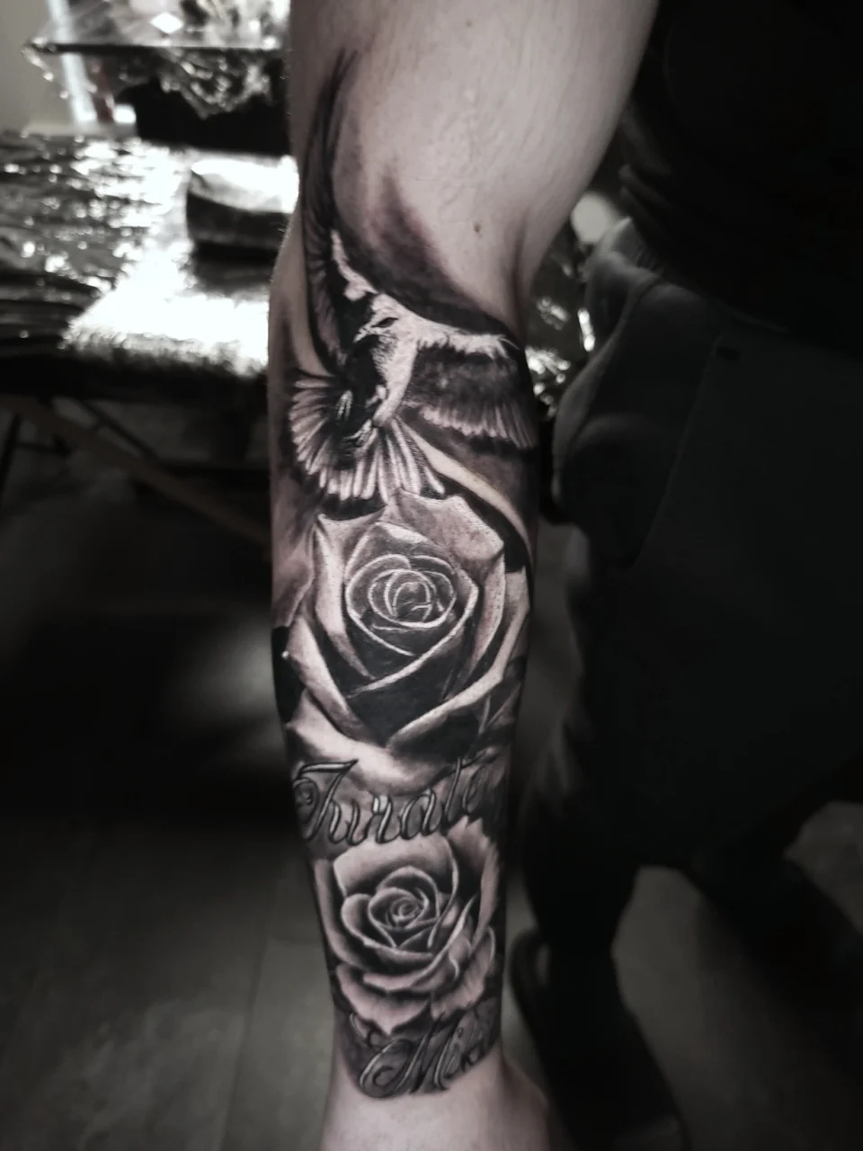 A black and grey tattoo on a forearm features a detailed rose, a flying bird, and script lettering. The tattoo covers most of the forearm and has realistic shading and intricate details.