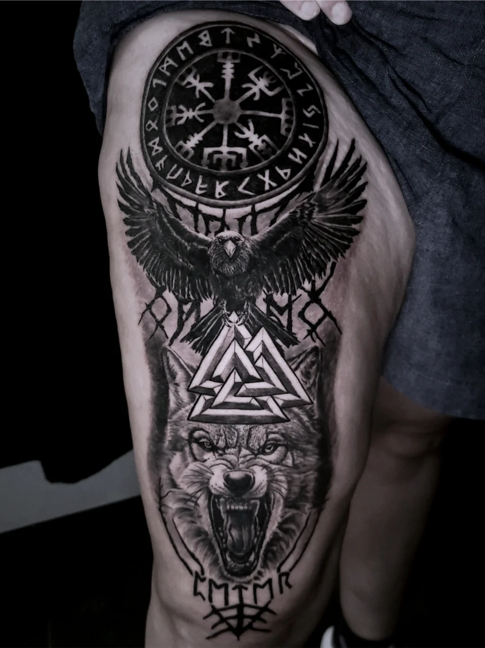 Black and grey thigh tattoo featuring Norse runes, a helm of awe symbol, a raven with outstretched wings, a Valknut symbol, a snarling wolf, and additional runes at the bottom.