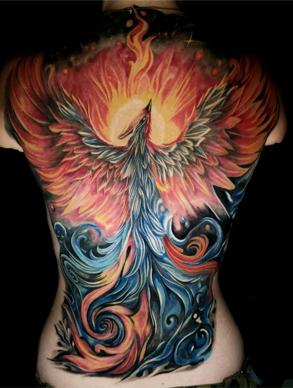 A vibrant tattoo of a phoenix with fiery red, orange, and yellow wings and blue swirling tail feathers covers a persons entire back, symbolizing rebirth and rising from flames.