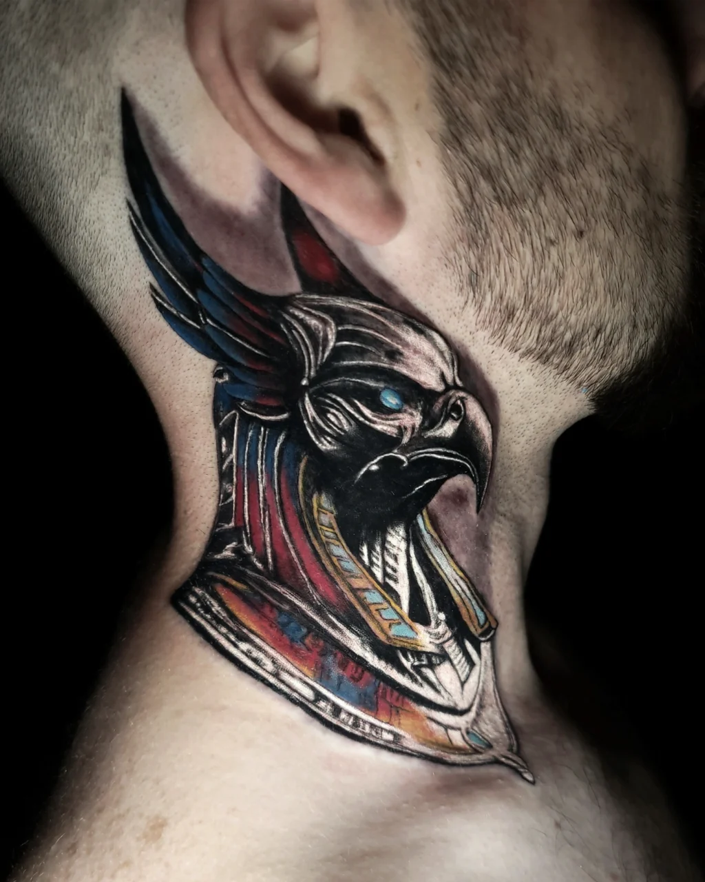 A detailed tattoo of an Egyptian-style falcon-headed deity with a colorful headdress is inked on the side of a persons neck, featuring bold black, red, and blue accents.