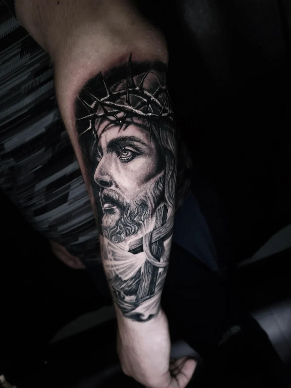 Black and grey tattoo of Jesus wearing a crown of thorns on a persons forearm, featuring detailed shading and realistic facial features.