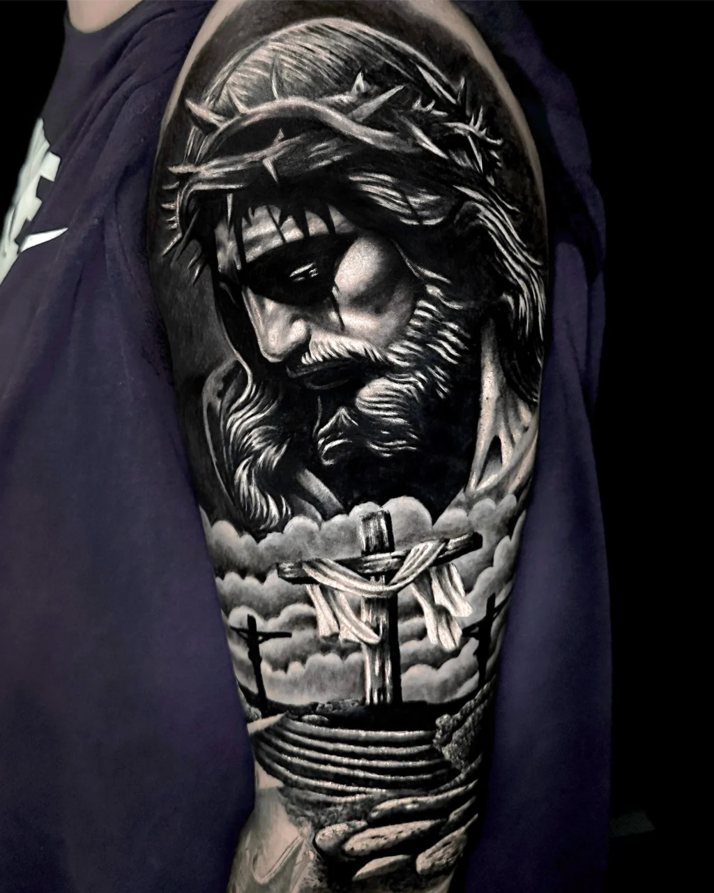 Realistic black and grey tattoo on an upper arm depicting Jesus with a crown of thorns above a cross draped with cloth, set against clouds and a dramatic sky.