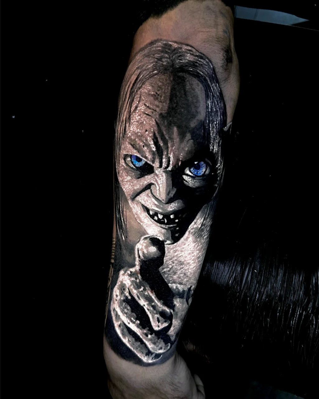 A realistic tattoo of a menacing character with long hair, sharp teeth, and glowing blue eyes, holding a small object in its hand, depicted on a persons forearm.