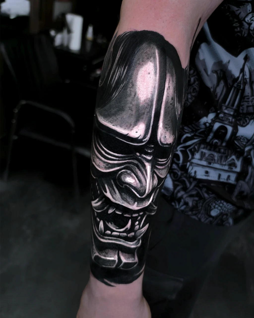 A close-up of a forearm tattoo depicting a detailed, black and grey Japanese samurai mask with fierce, expressive features and sharp teeth. The background is slightly blurred, focusing on the tattoo art.