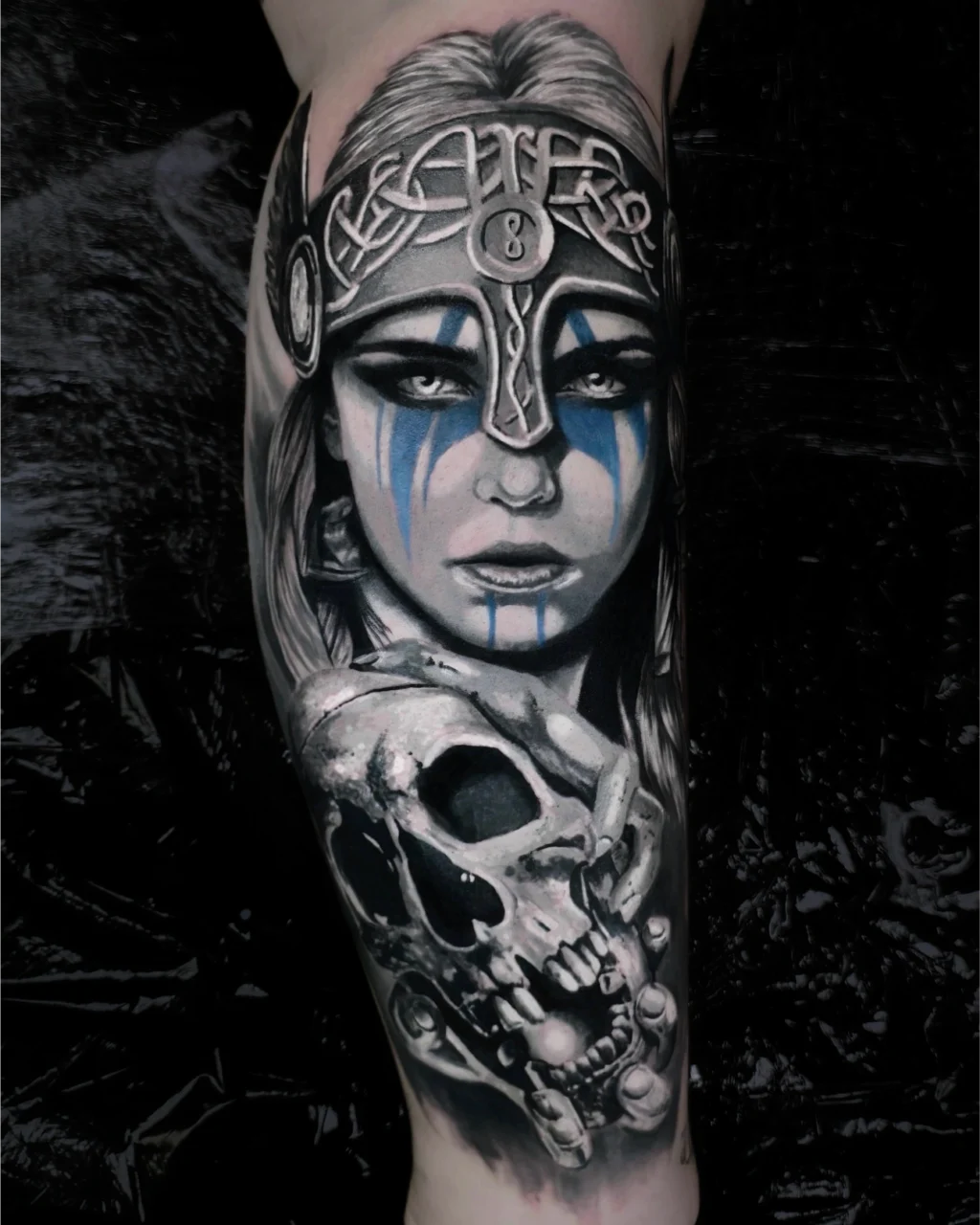 A realistic black and grey tattoo of a Viking woman with blue war paint on her face, wearing a helmet with Norse designs, above a detailed skull adorned with beads and trinkets.