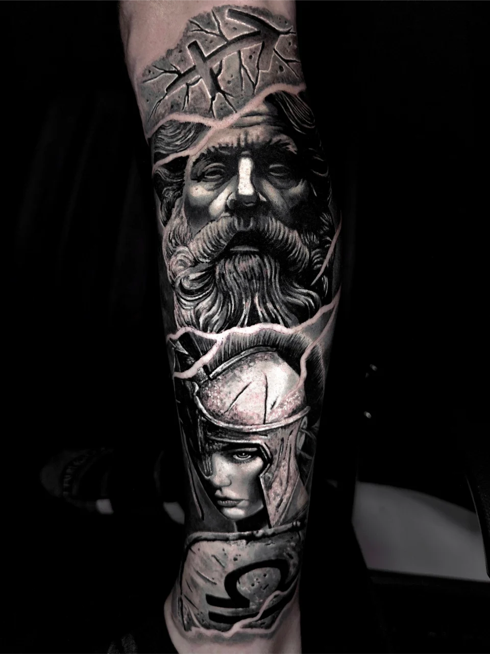 Realistic black and grey tattoo of two ancient figures: an older bearded man on top, and a helmeted warrior below, both surrounded by stone and armor elements, inked on a persons forearm.