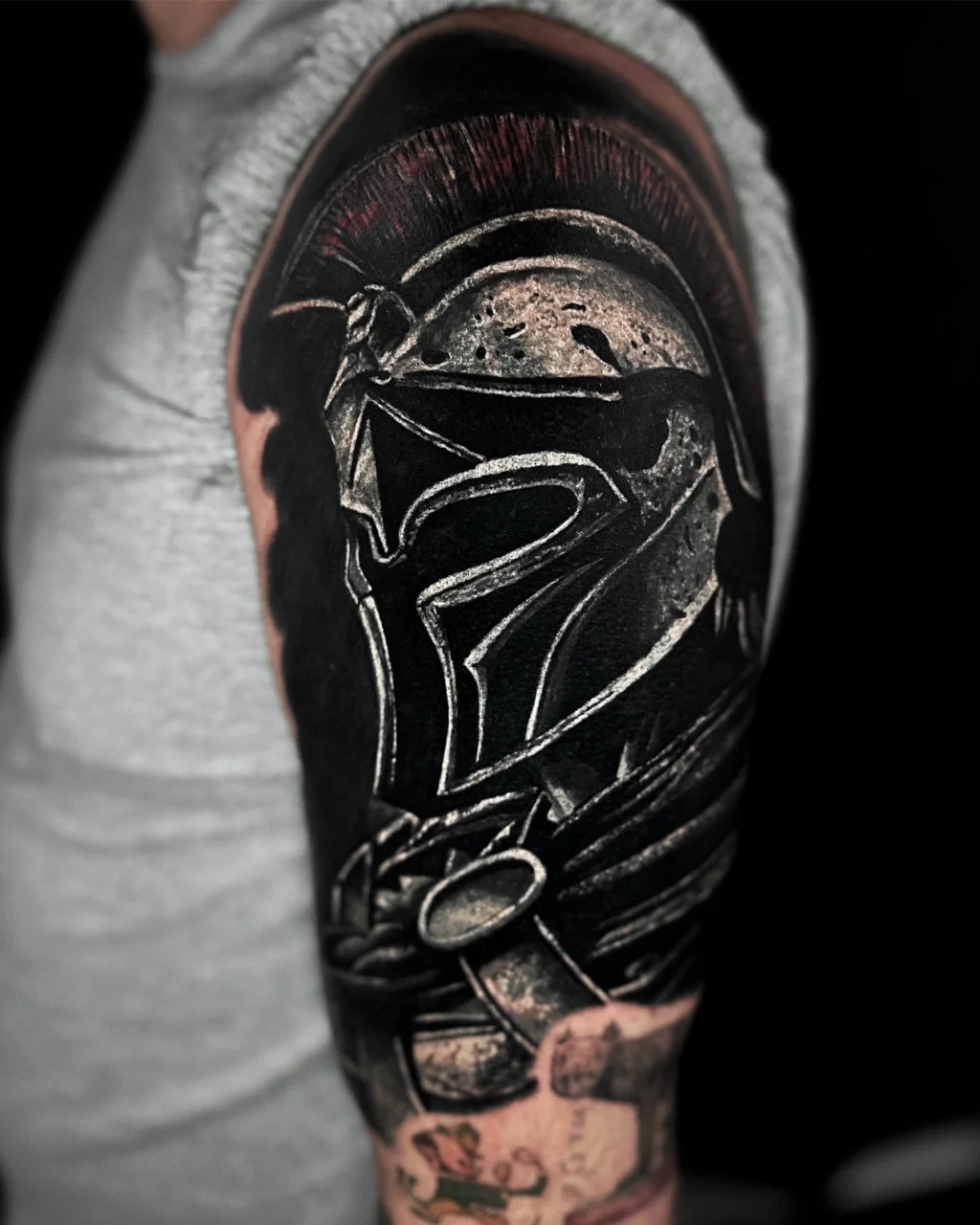 A detailed tattoo of a Spartan helmet with a red plume, covering the upper arm of a person wearing a grey sleeveless shirt. The design features strong black and white shading for a dramatic effect.