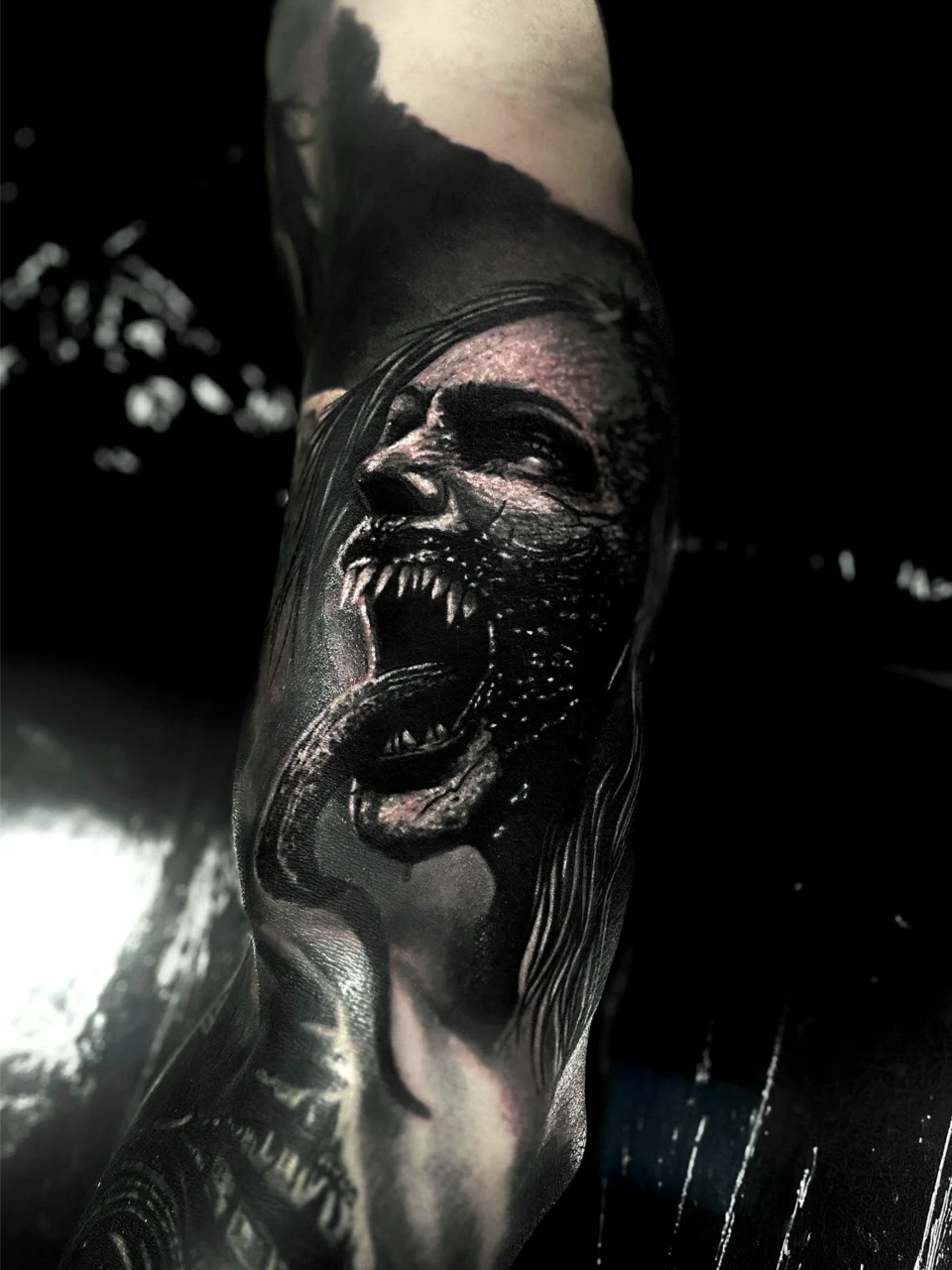 A realistic black and grey tattoo on an arm depicts a fierce, snarling face with sharp fangs and a long tongue, blending into dark shadows and detailed hair.
