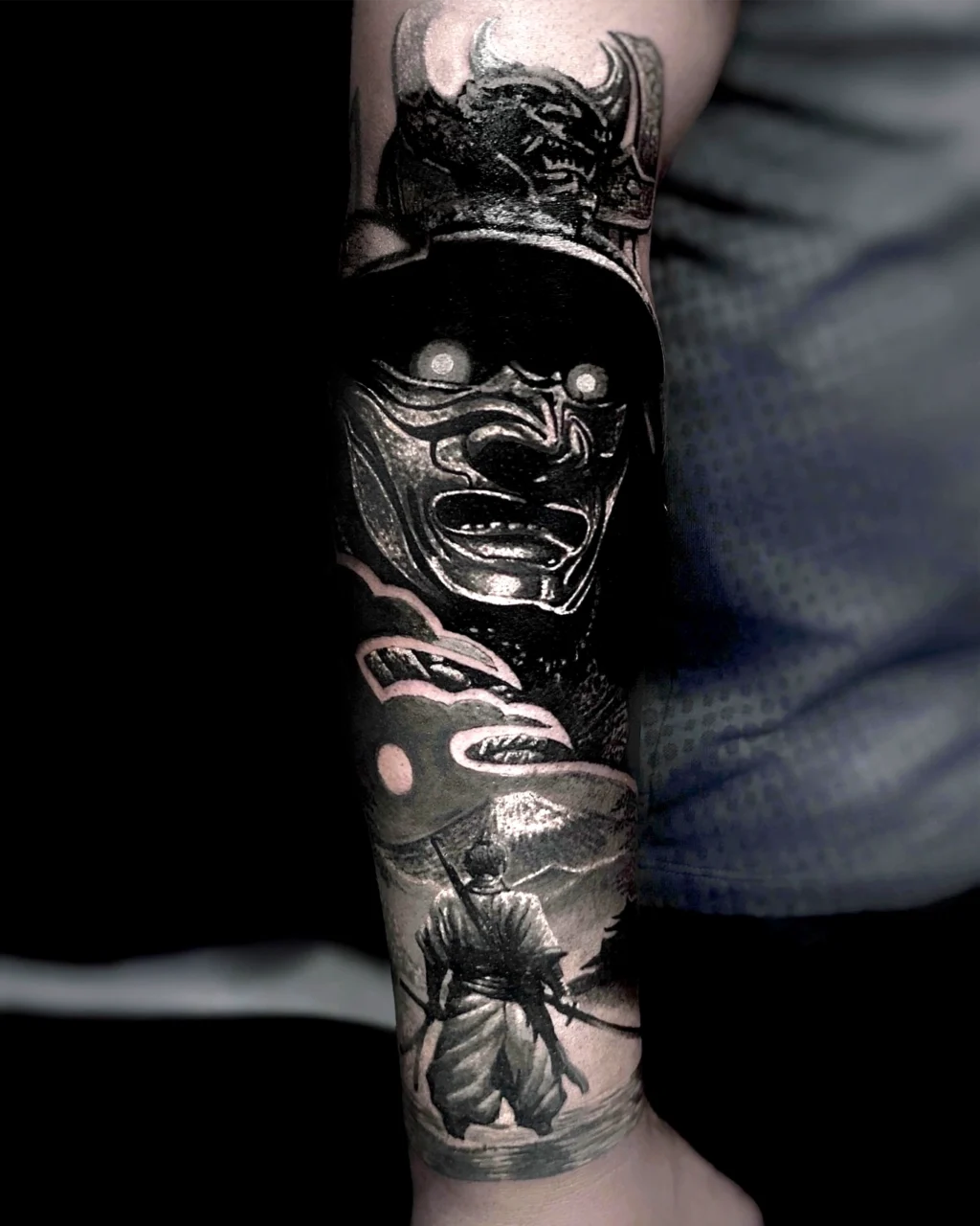 A detailed black and gray tattoo on an arm shows a glowing-eyed samurai mask above a scene of a lone samurai with two swords standing before mountains and clouds.