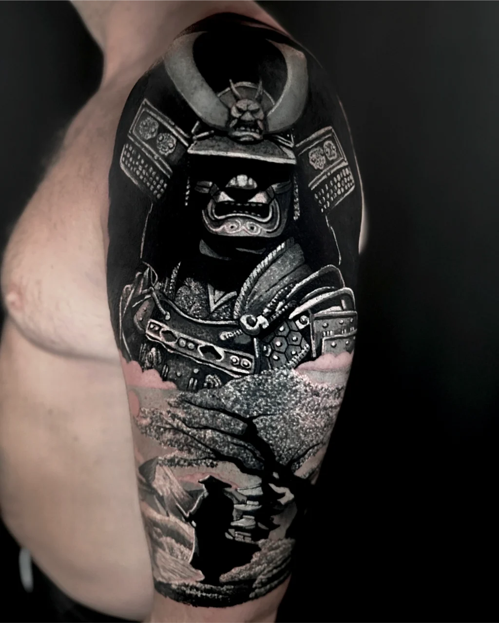 A detailed black and gray tattoo on an upper arm features a samurai in traditional armor above a landscape with a lone figure standing beneath a tree. The tattoo covers the shoulder and upper arm.