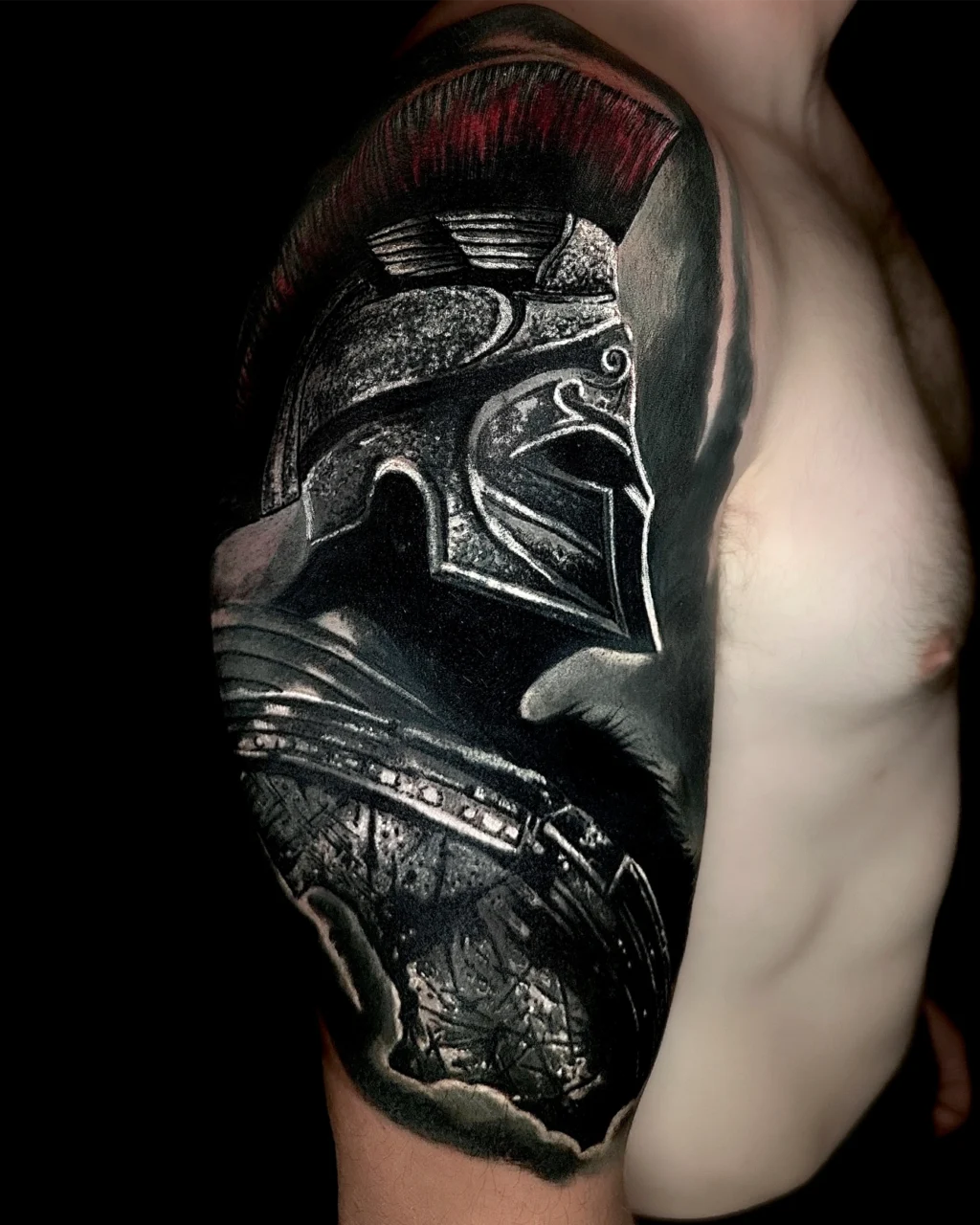 A detailed tattoo of a Roman or Greek helmeted warrior covers the upper arm and shoulder of a shirtless person, featuring black, grey, and red ink with realistic shading and intricate armor details.