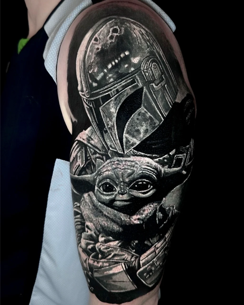 A detailed black and gray tattoo on an upper arm depicts the Mandalorian and Grogu (Baby Yoda) from the Star Wars series, with intricate shading and realistic features.