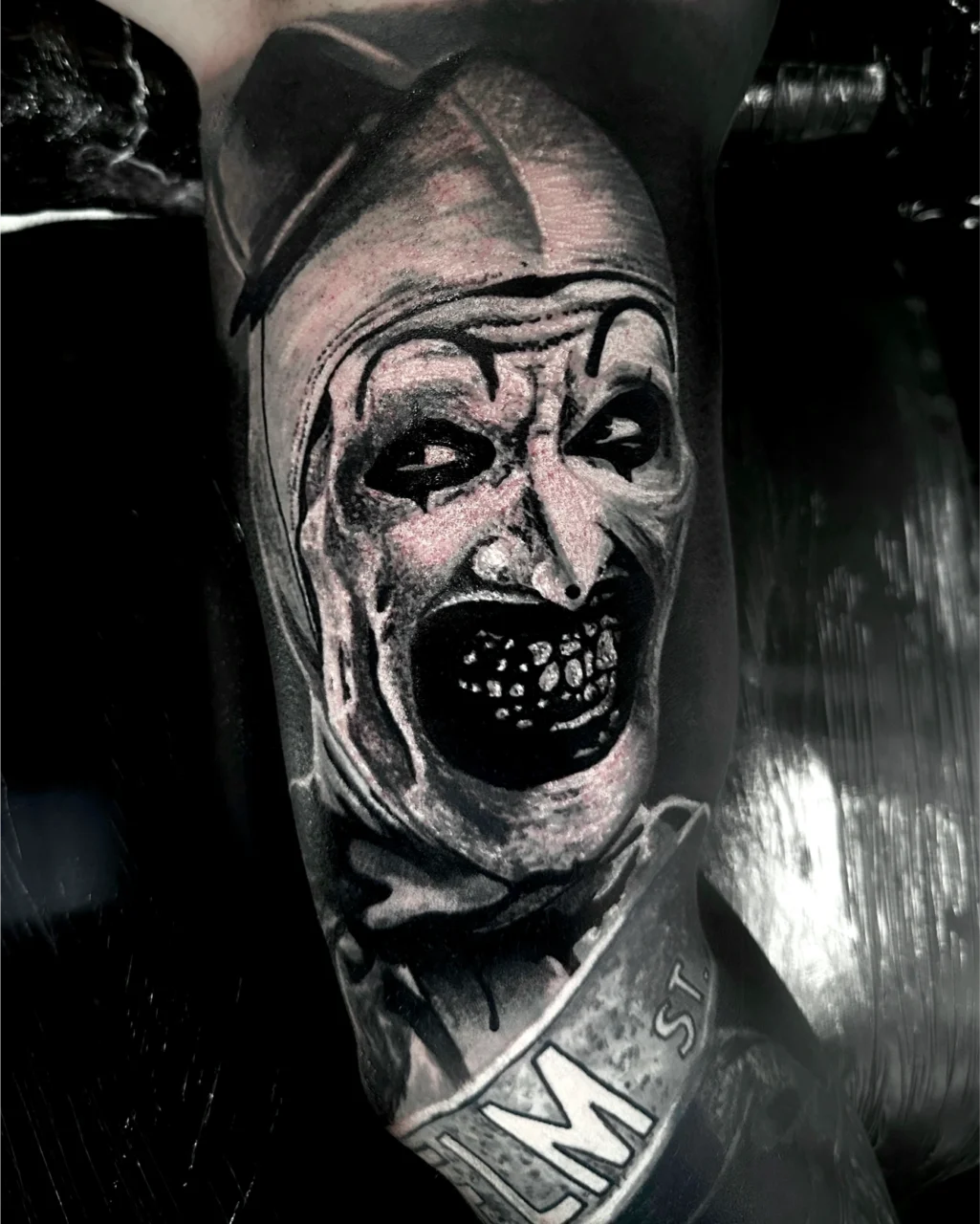A black and gray realistic tattoo of a sinister clown with a wide, open mouth showing dark, jagged teeth and exaggerated facial features. The lower part has partial text reading LM ST.