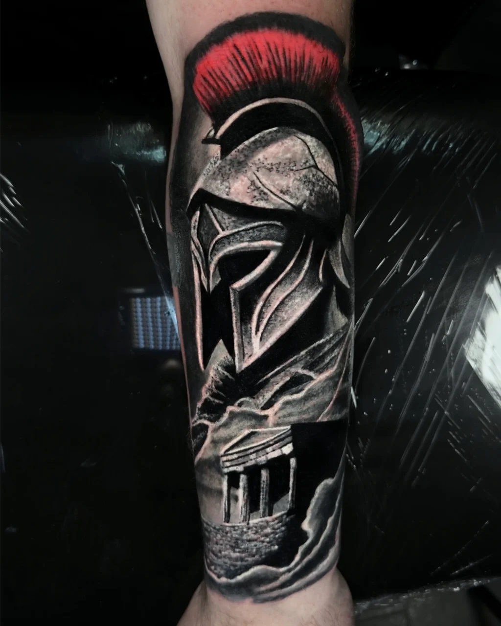 A realistic tattoo of a Greek or Roman warrior helmet with a red crest, above an ancient temple surrounded by clouds, inked in black, gray, and red on a persons forearm.