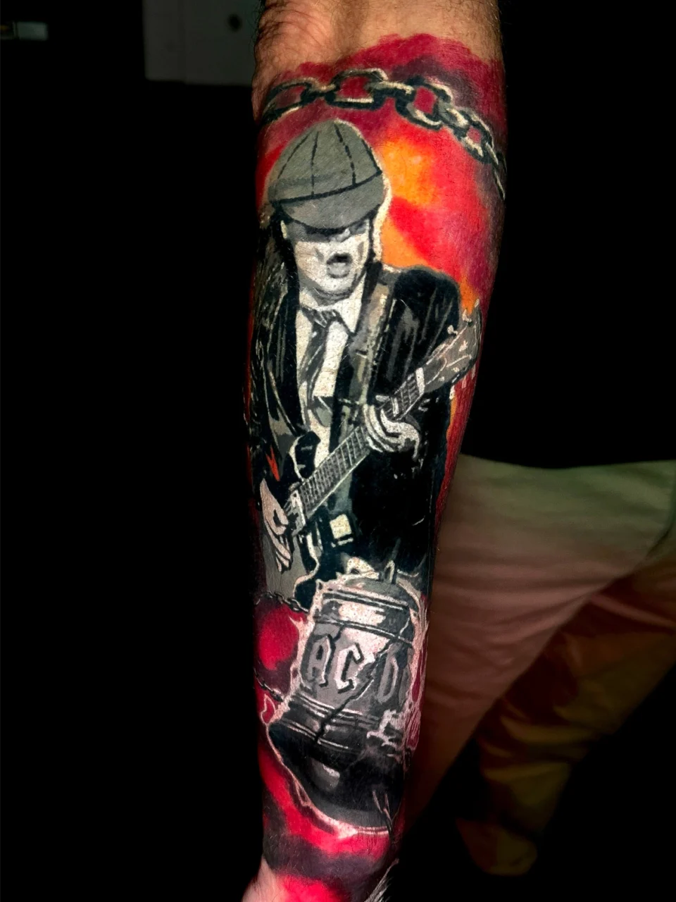 A tattoo on a forearm featuring a guitarist in a school uniform and cap, playing an electric guitar, with a red and black background and the letters ACDC on a stylized bell below.