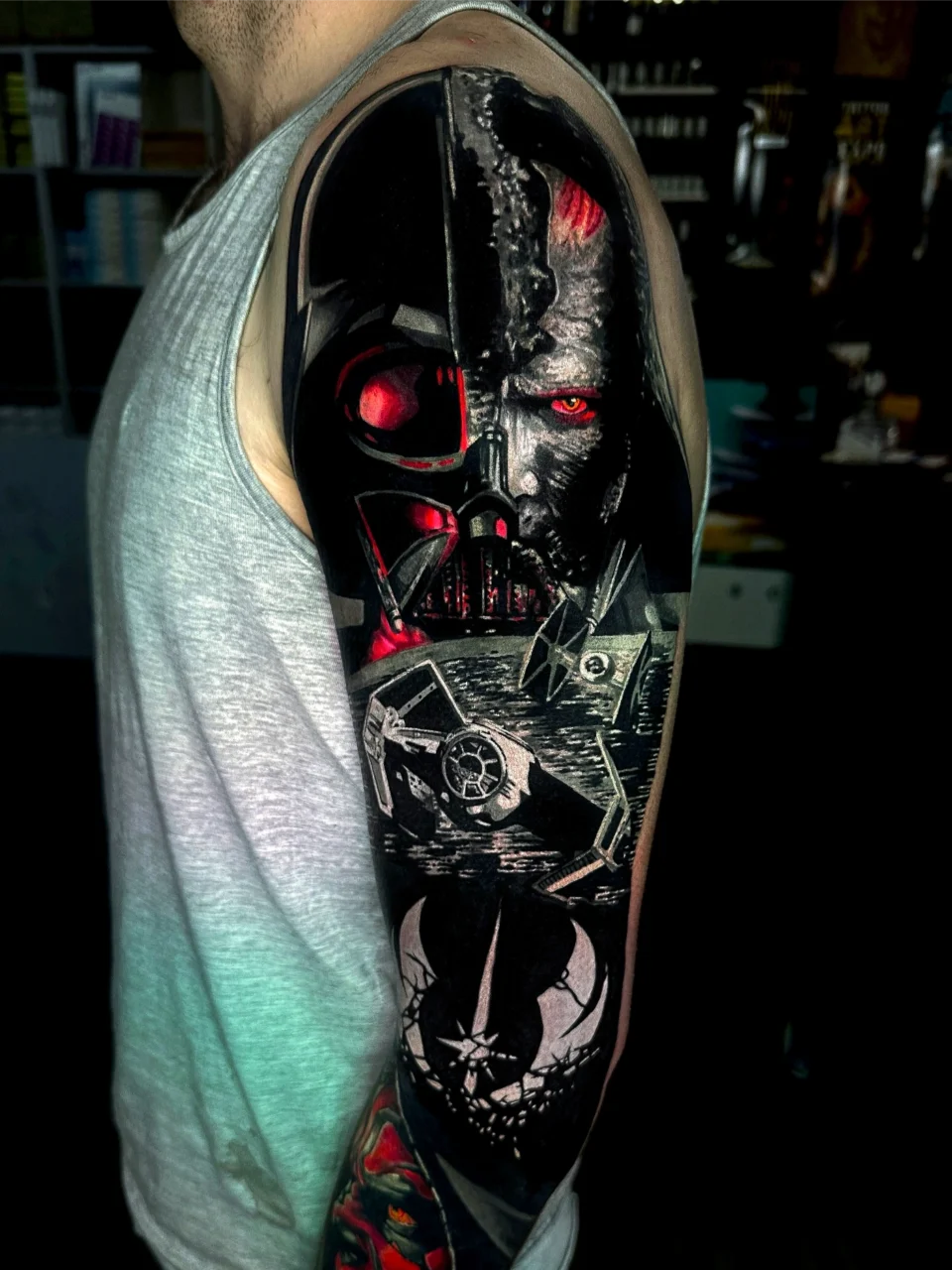 A person in a gray tank top shows a detailed Star Wars-themed tattoo sleeve featuring Darth Vader’s face, TIE fighters, and the Empire emblem in vibrant black, red, and white ink.