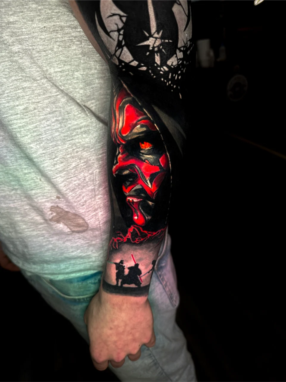 A person’s forearm tattoo featuring a vivid red and black portrait of Darth Maul from Star Wars, above a silhouette of two figures with lightsabers in a duel. The tattoo extends across the arms length.