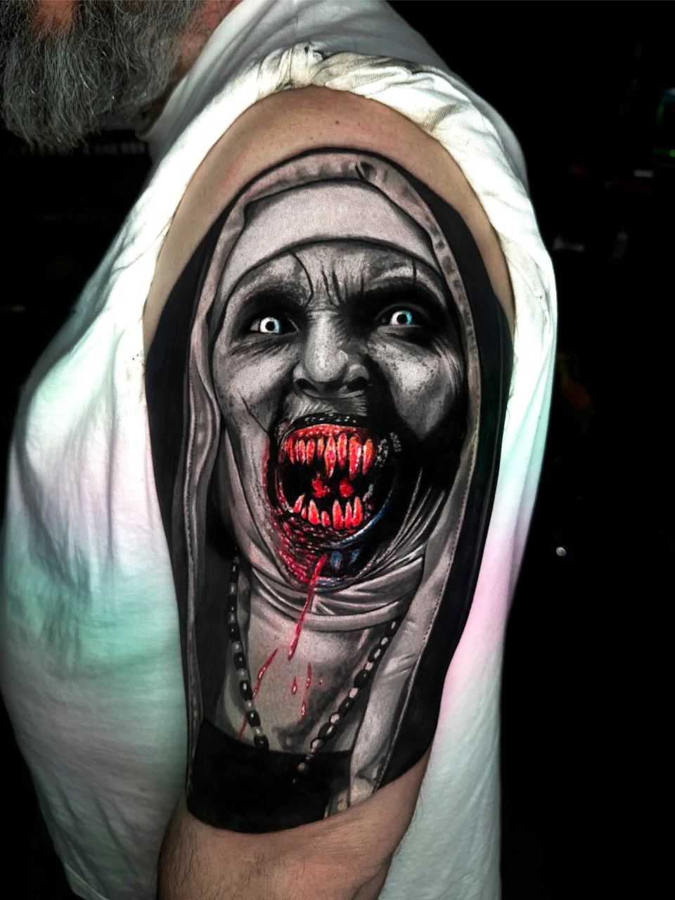 A tattoo on an upper arm depicts a demonic nun with glowing blue eyes, sharp bloody teeth, and an open mouth dripping blood. The image is highly detailed, with dark shading and horror elements.